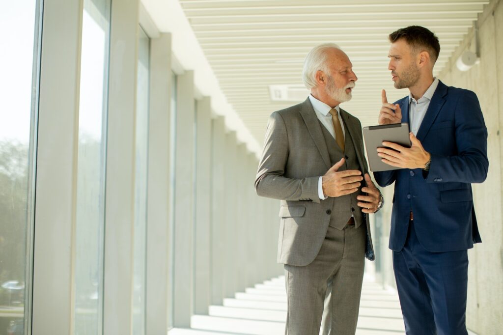 Young and a senior businessman walk down an office hallway, deep in conversation